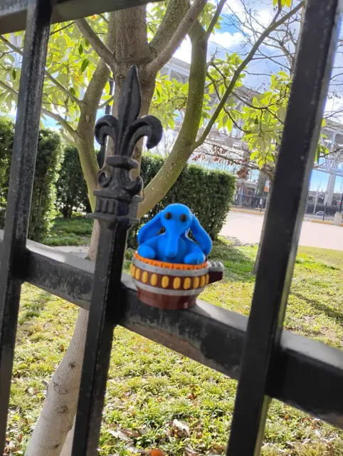 A small blue Max Rebo figure perched on a black iron fence, with a tree and a lawn behind it.