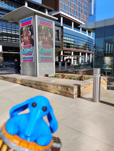 A small blue Max Rebo figure in the foreground of a paved plaza, with a poster kiosk and glass buildings behind it.